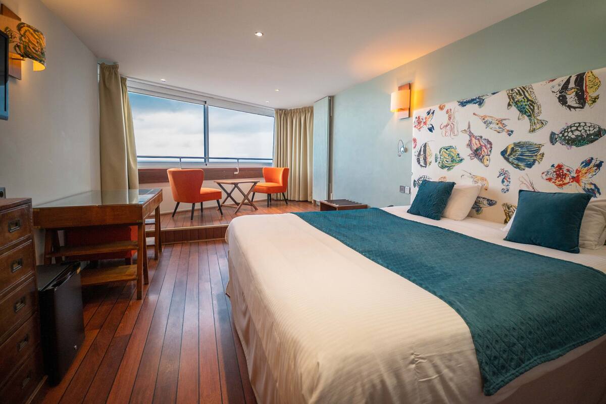 Habitación de hotel con una cama grande, un escritorio y una silla. Hay una ventana con cortinas, una pared con un diseño de peces y vistas al cielo.