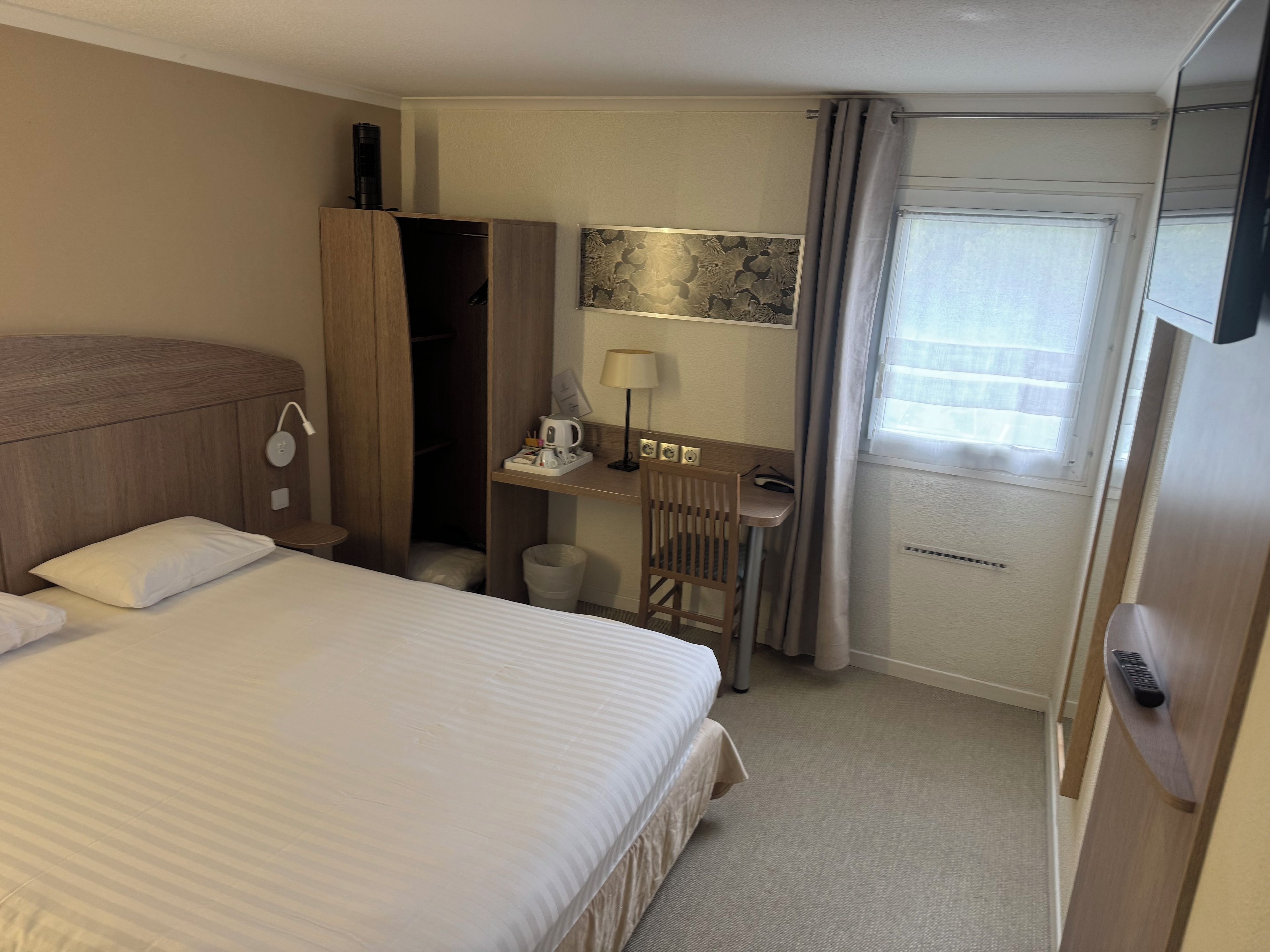 Hotellihuone, jossa on sänky, työpöytä, tuoli, lamppu, kahvinkeitin ja ikkuna, jossa on verhot.
