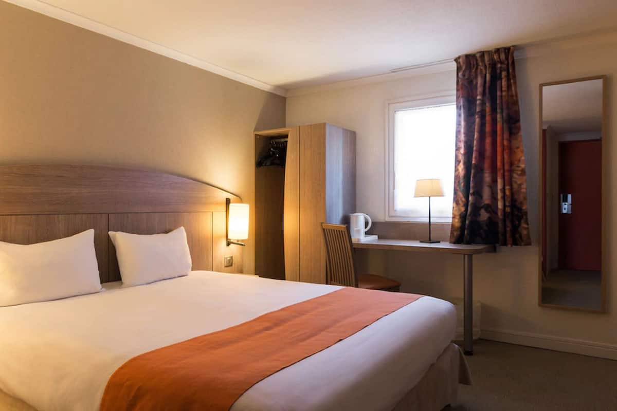 Une chambre d’hôtel avec un grand lit, un bureau avec une chaise, une armoire et une fenêtre avec des rideaux.