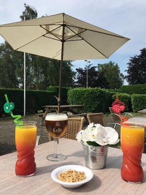 An outdoor dining area with a table set for two, featuring a glass of beer, two fruit juices, a bowl of nuts, and a metal cup. - The Originals City, Hôtel Le Gayant (Douai)