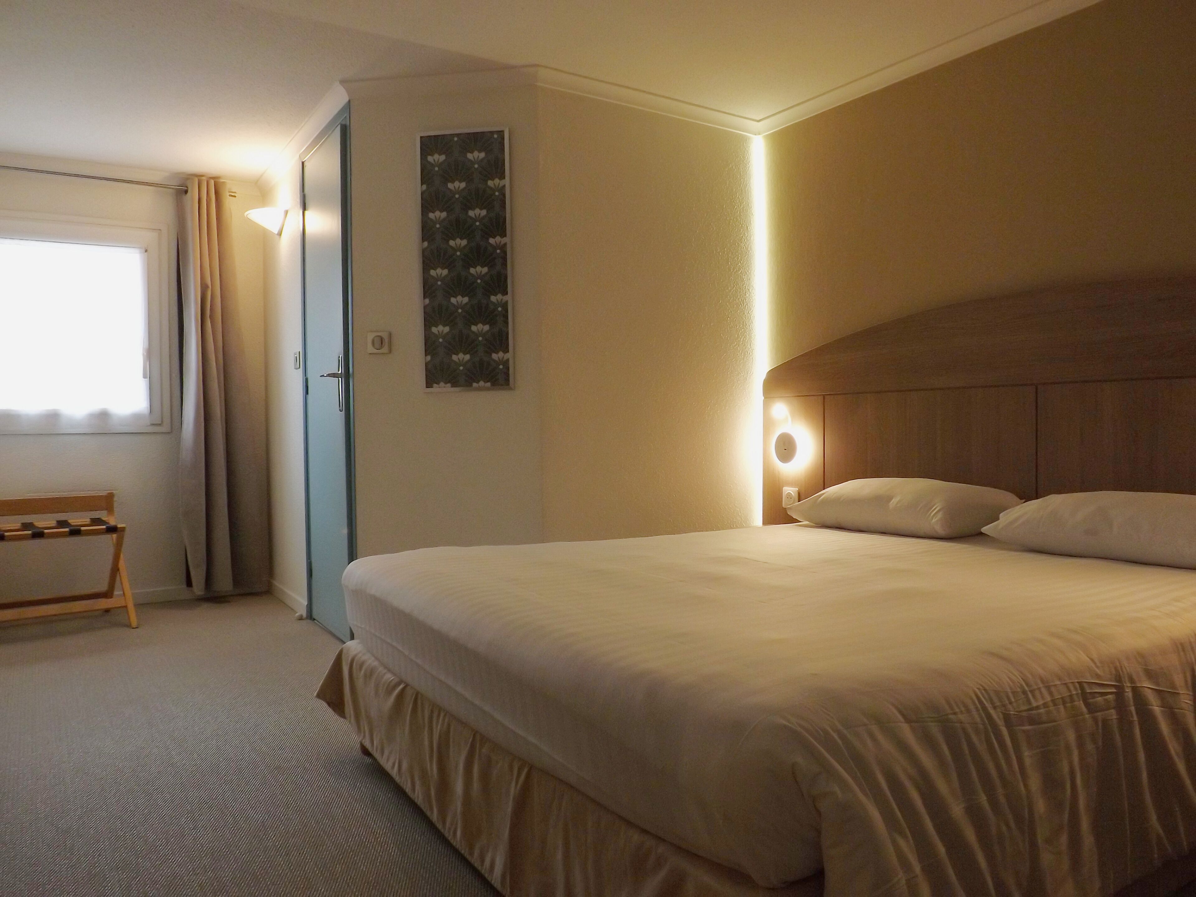 Een hotelkamer met een bed, nachtkastje, een wandlamp en een deur.