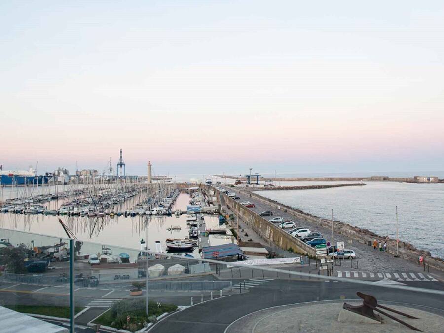A marina with boats docked, a large ship in the background, and a road leading to the water.