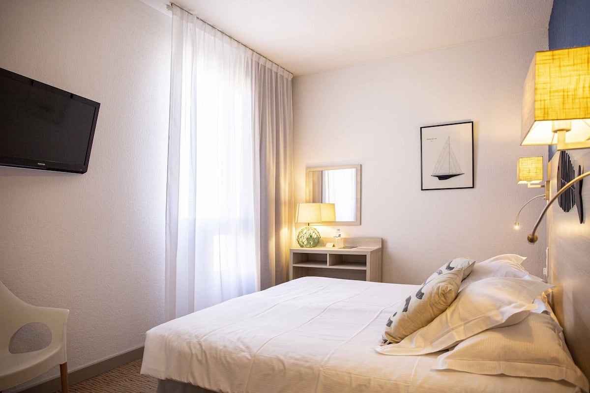 Quarto de hotel com cama, televisão fixada na parede, secretária com luminária, espelho e janela com cortinas.
