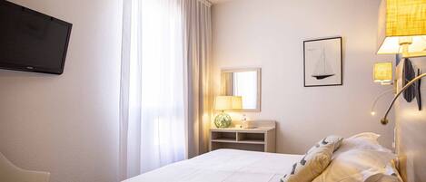 Une chambre d’hôtel avec un lit, un téléviseur fixé au mur, un bureau avec une lampe, un miroir et une fenêtre avec des rideaux.