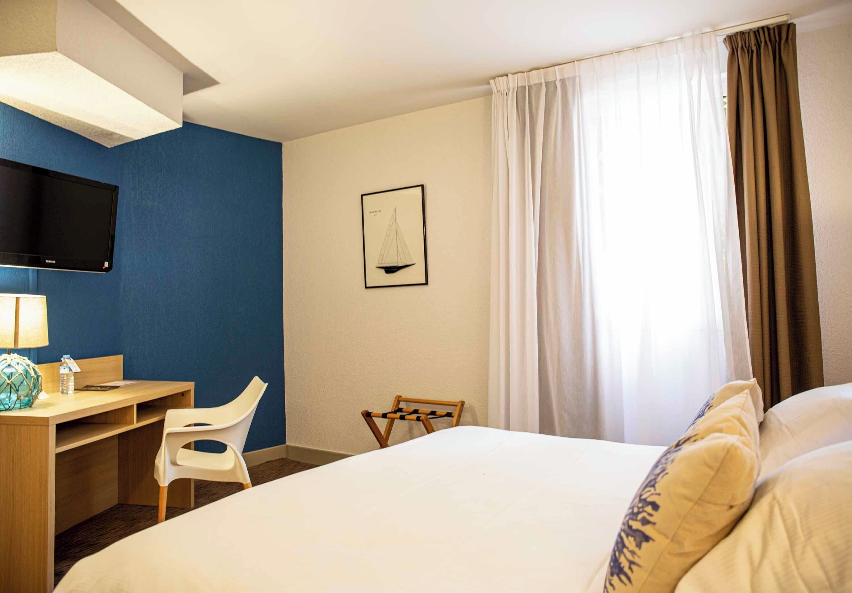 A hotel room with a bed, a desk with a chair, a television, and a window with curtains.