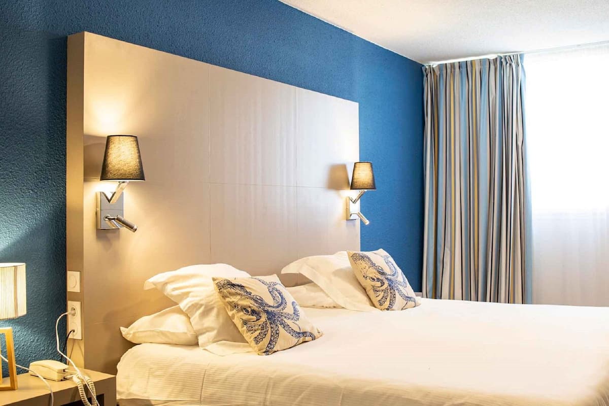 Quarto de hotel com uma cama grande, duas luminárias de cabeceira, um telefone e vista exterior através de uma janela com cortinas.