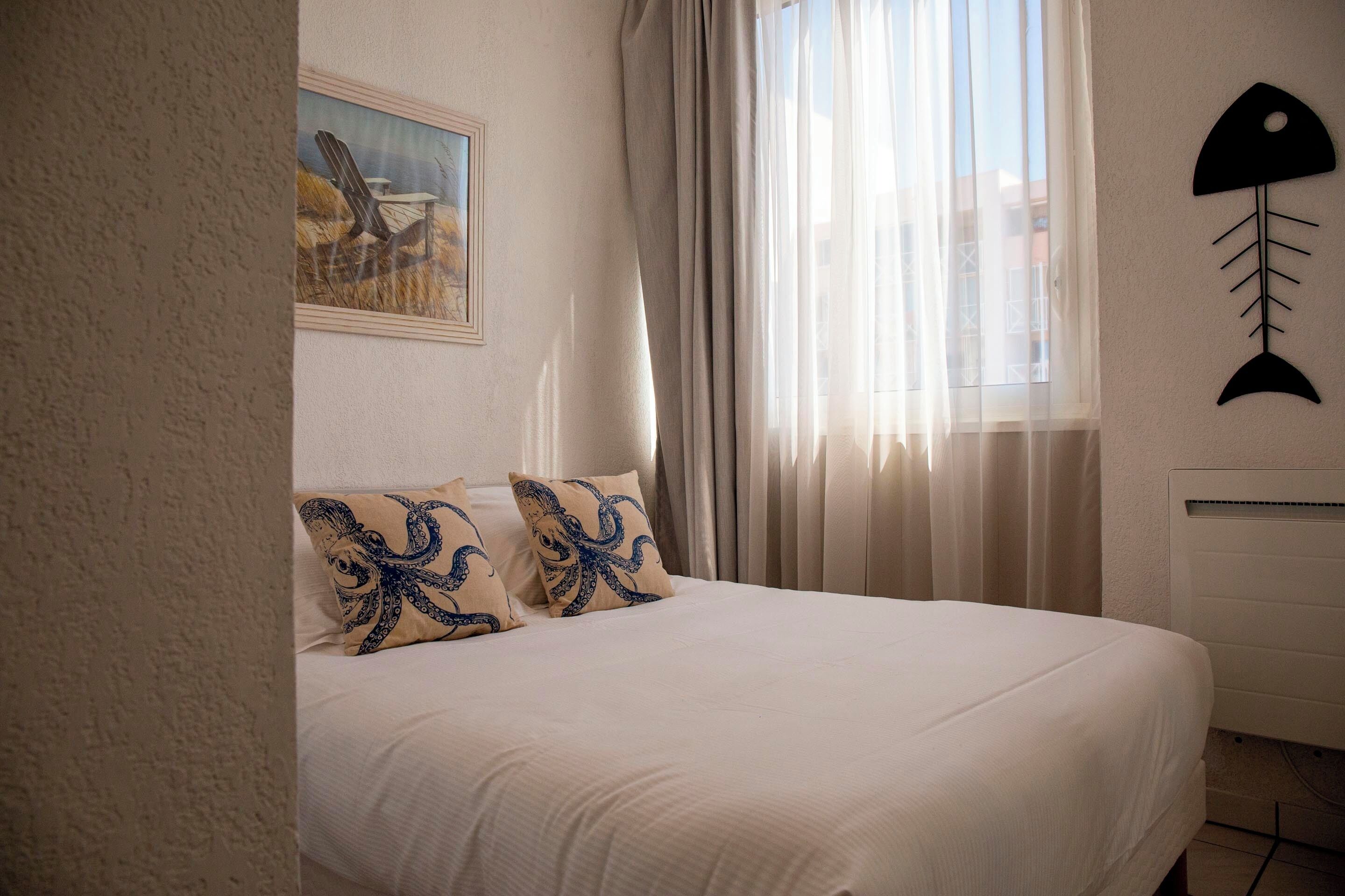 Una cama con ropa blanca, un cuadro de una playa y un adorno de pez en la pared.