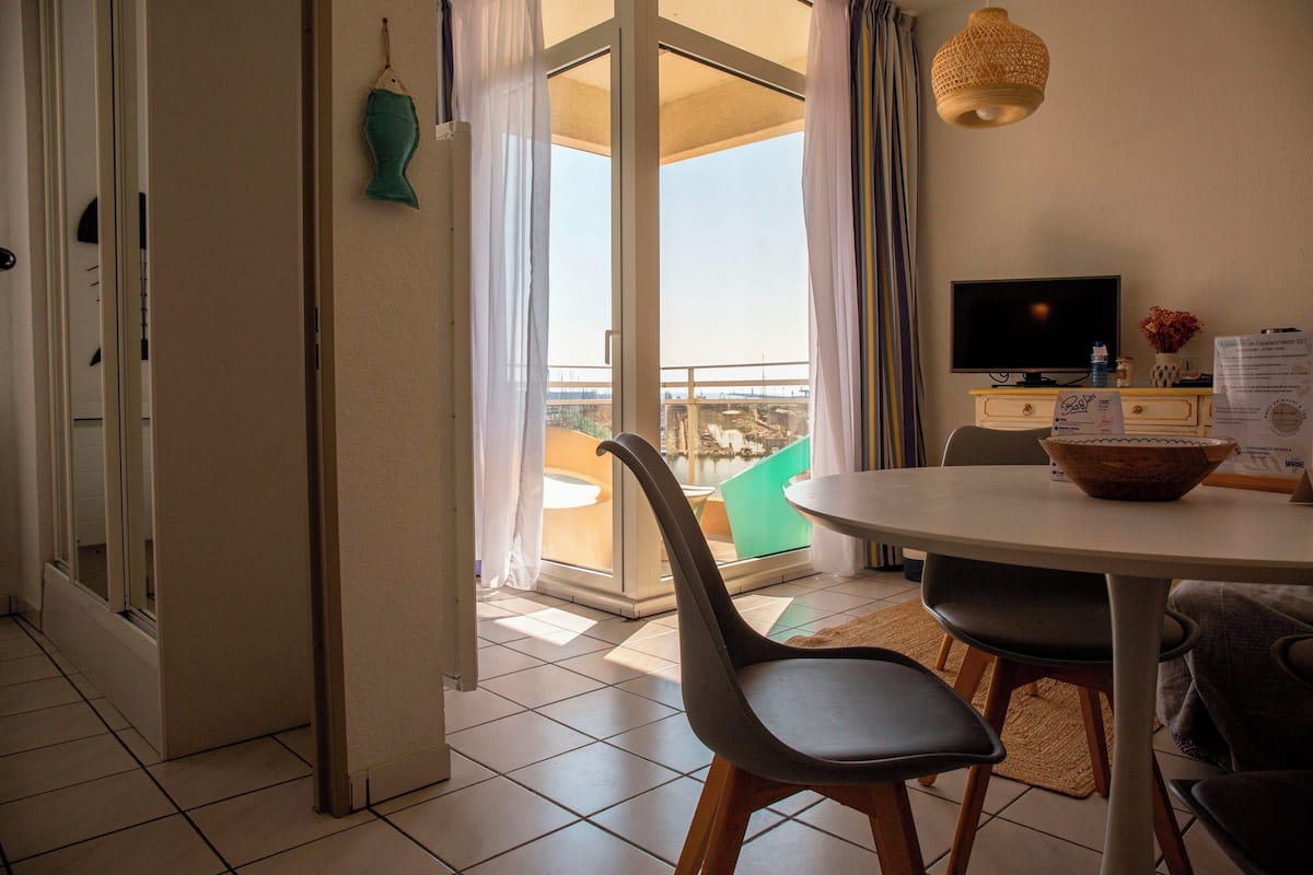 Quarto com mesa de jantar, cadeiras, televisão e vista para o exterior.