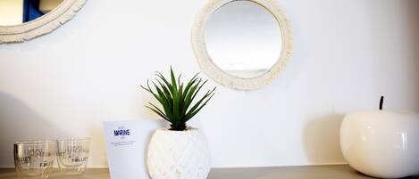 Pared blanca con tres espejos redondos y un jarrón blanco con forma de piña que contiene una planta. Hay dos vasos y una tarjeta en la encimera.