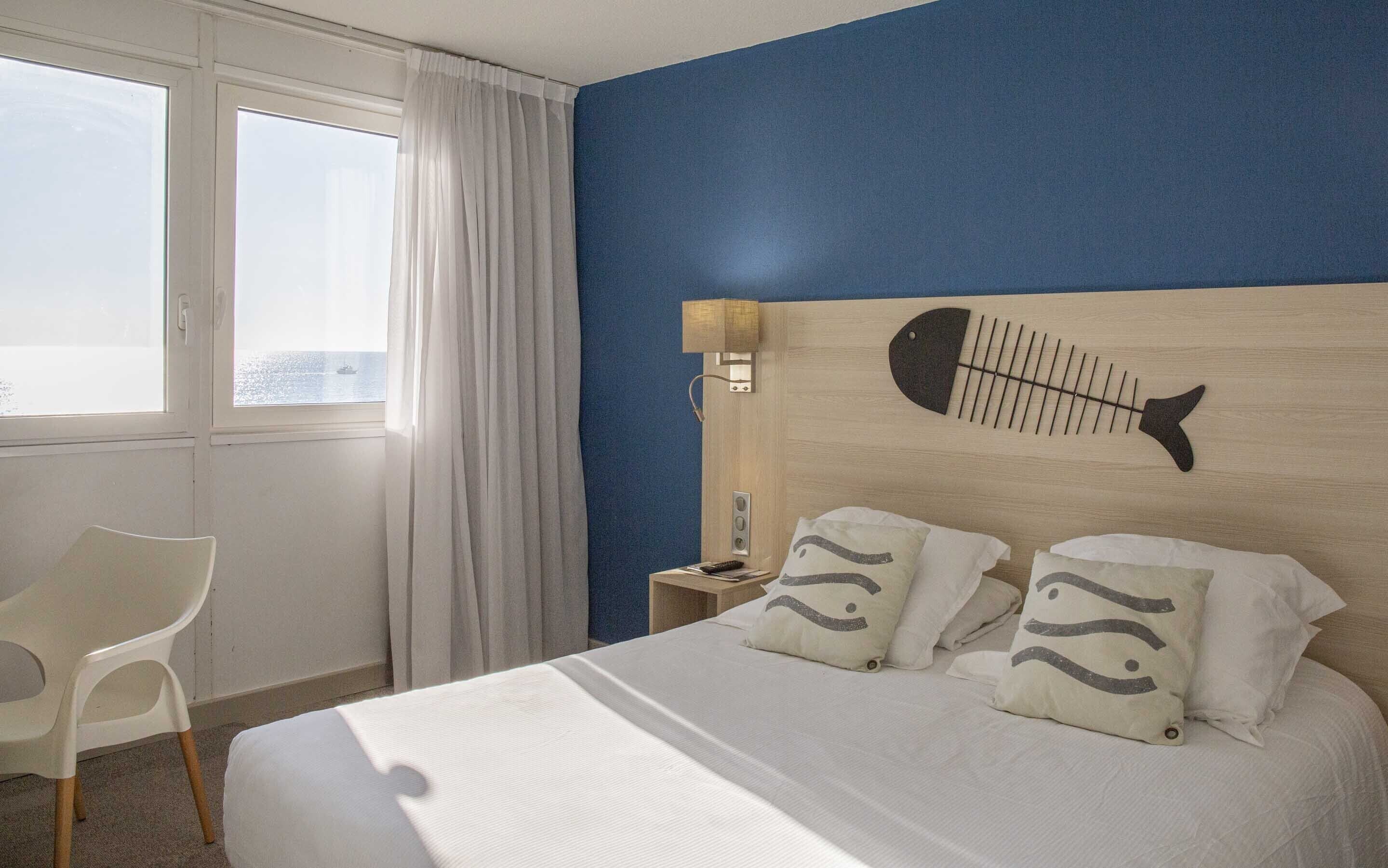 Habitación de hotel con cama, mesita de noche, lámpara, silla y vista al mar.