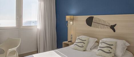 Habitación de hotel con cama, mesita de noche, lámpara, silla y vista al mar.