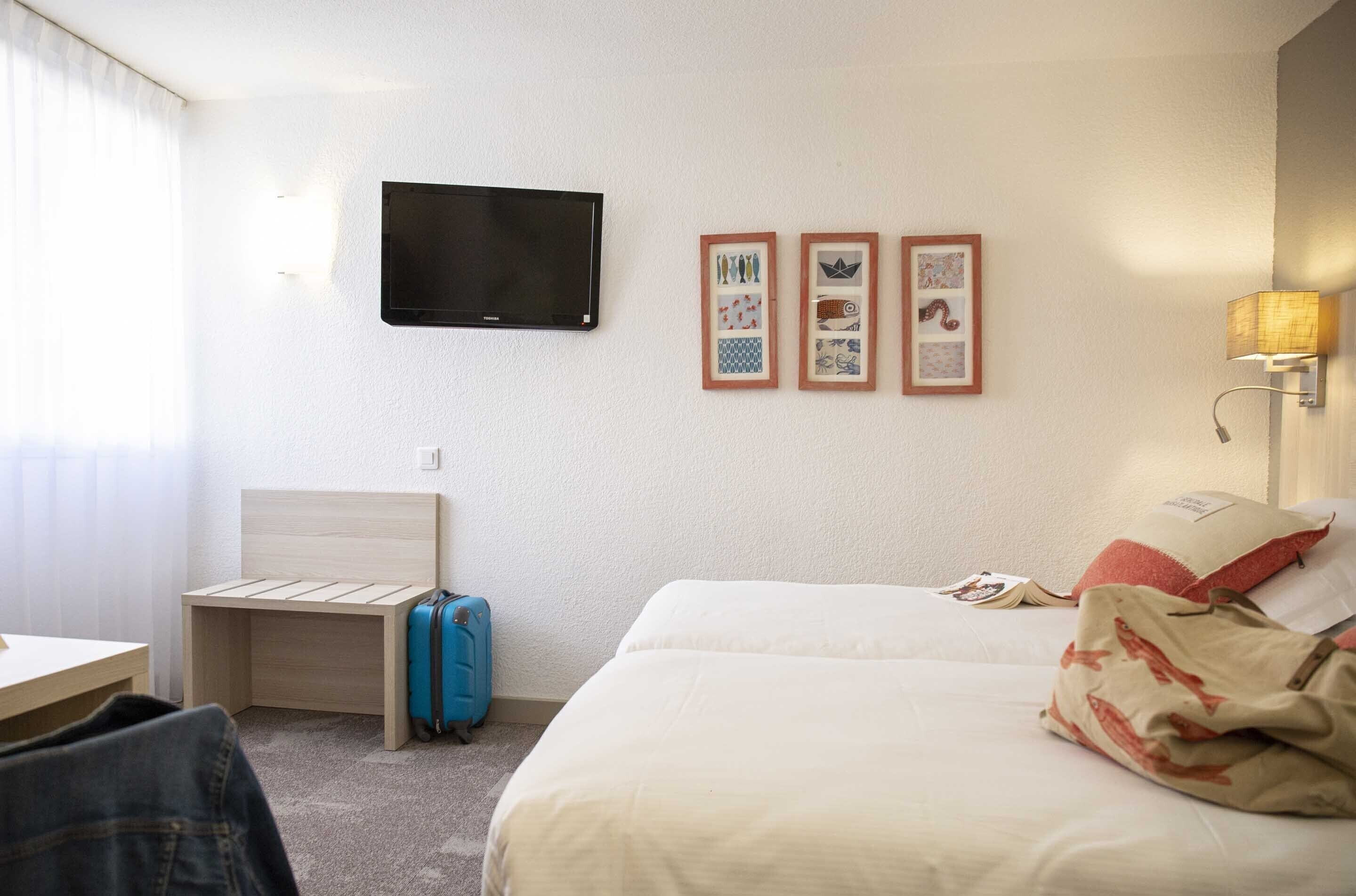 A hotel room with a bed, a desk, a chair, a TV, and framed pictures on the wall.