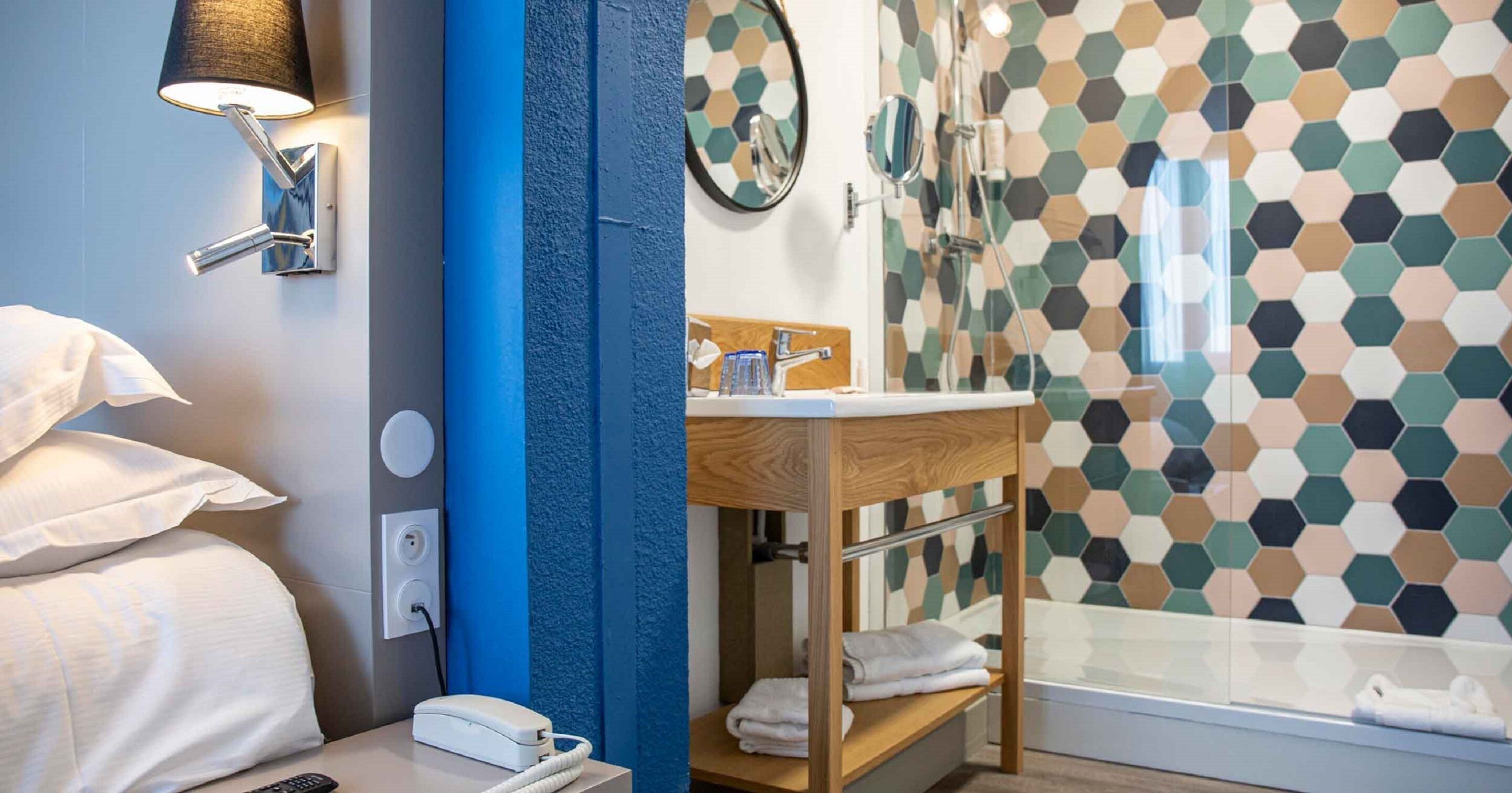A hotel bathroom with a shower, a sink, a mirror, and a bedside table with a lamp.