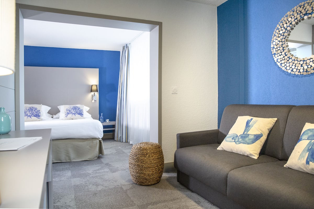 Quarto de hotel com cama, sofá, luminária, espelho e um banquinho de fibra sintética.