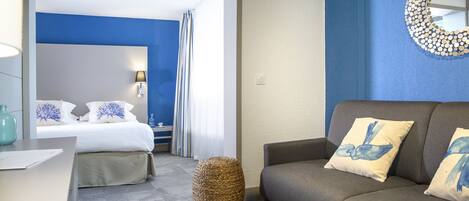 A hotel room with a bed, a sofa, a lamp, a mirror, and a woven stool.