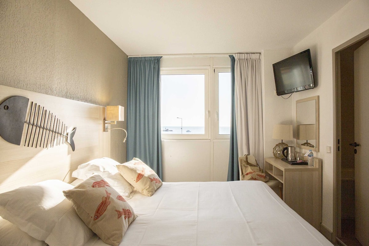 Quarto de hotel com cama, escrivaninha, televisão e janela com cortinas.