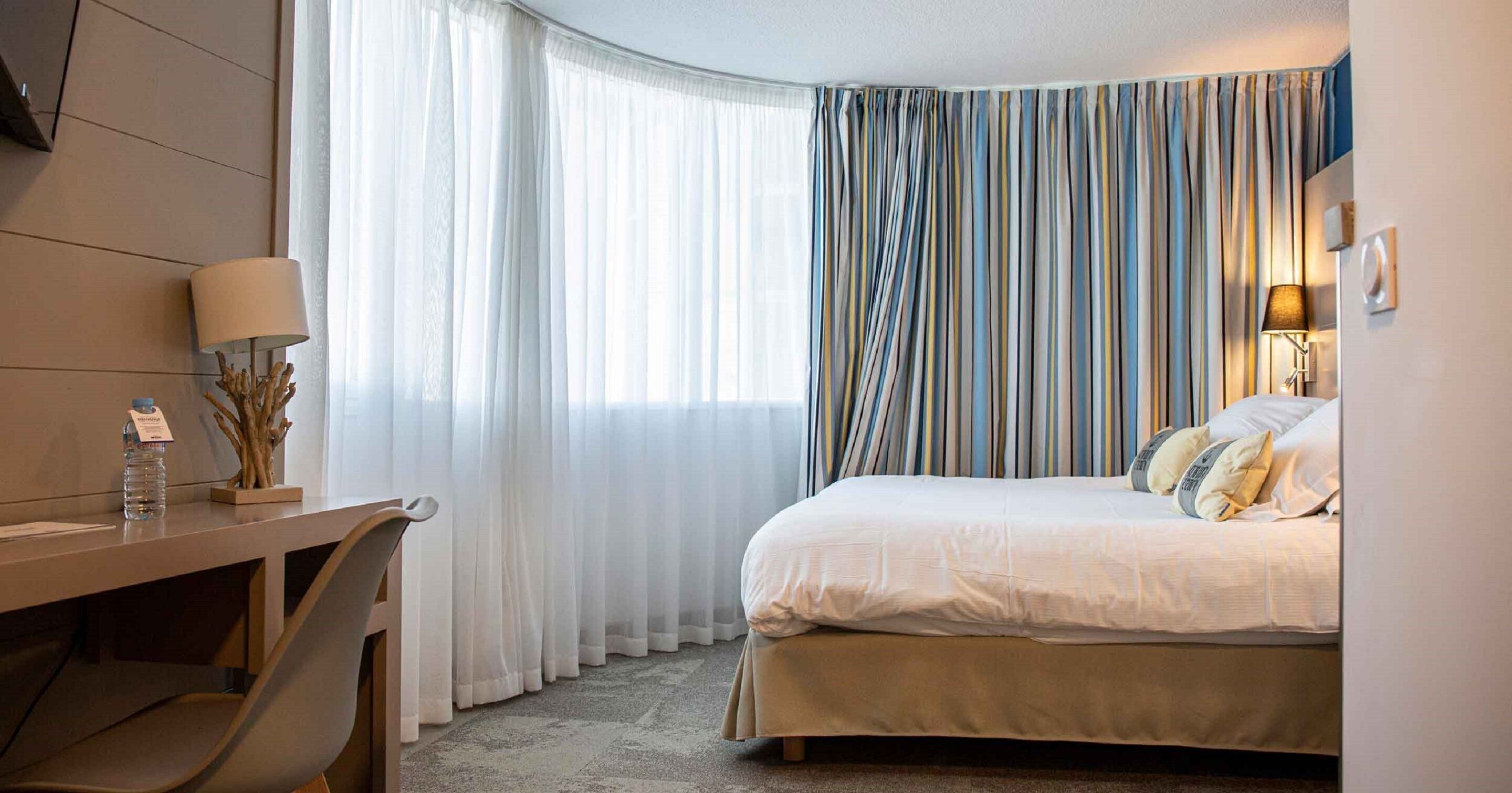 Een hotelkamer met een bed, een bureau, een stoel, een lamp en een raam met gordijnen.