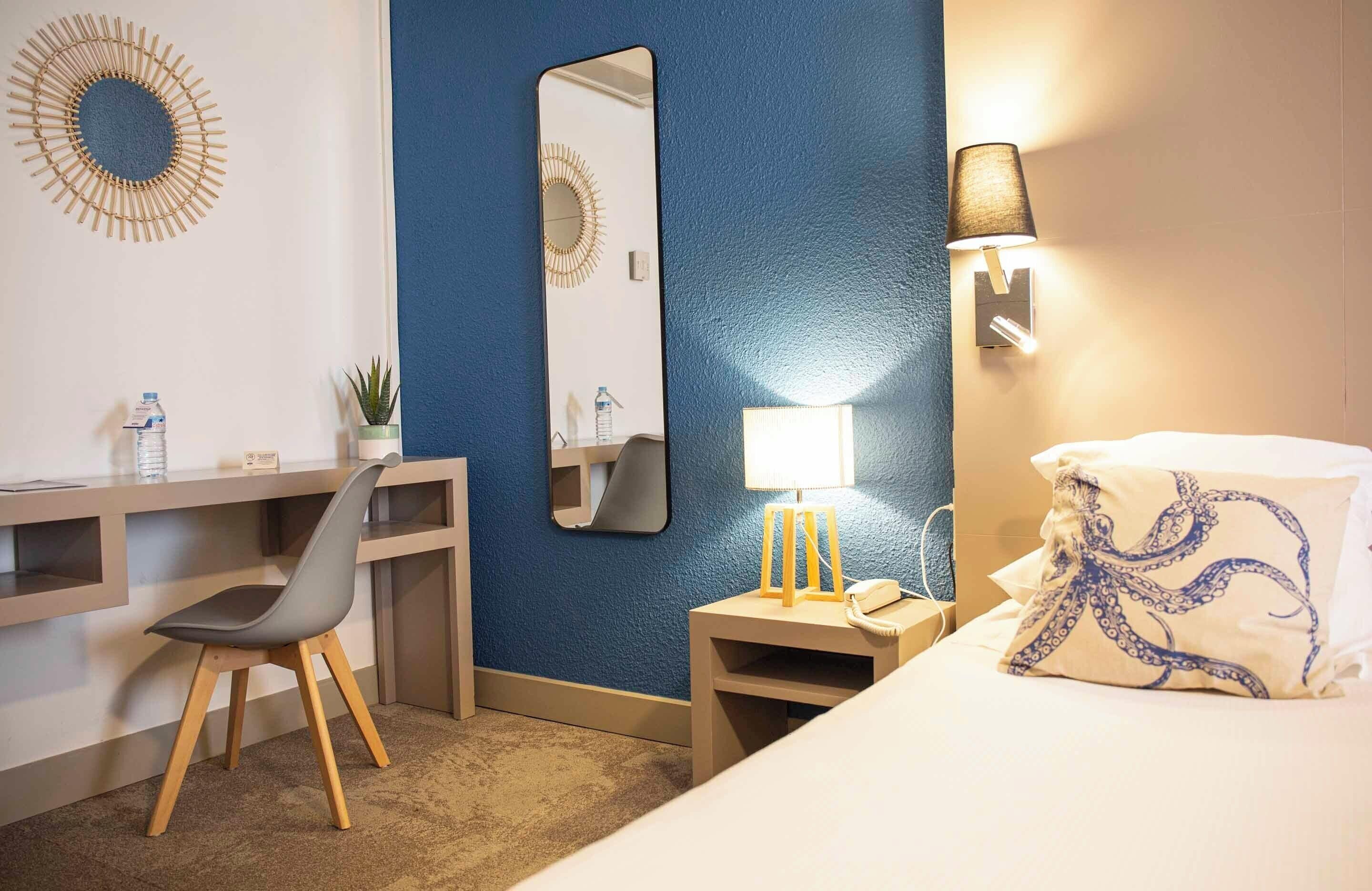 Hotellihuone, jossa on työpöytä, tuoli, sänky, yöpöytä, lamppu ja seinälle kiinnitetty peili.