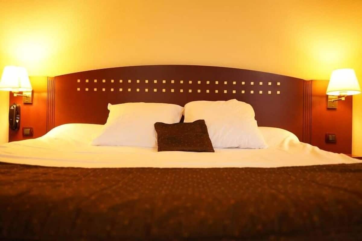 Una cama bien tendida con sábanas blancas y dos almohadas, cabecera oscura y dos lámparas adosadas a la pared a cada lado.