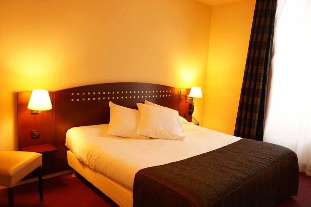 Habitación de hotel con una cama grande, dos mesitas de noche con lámparas, una silla y un banco de color oscuro.