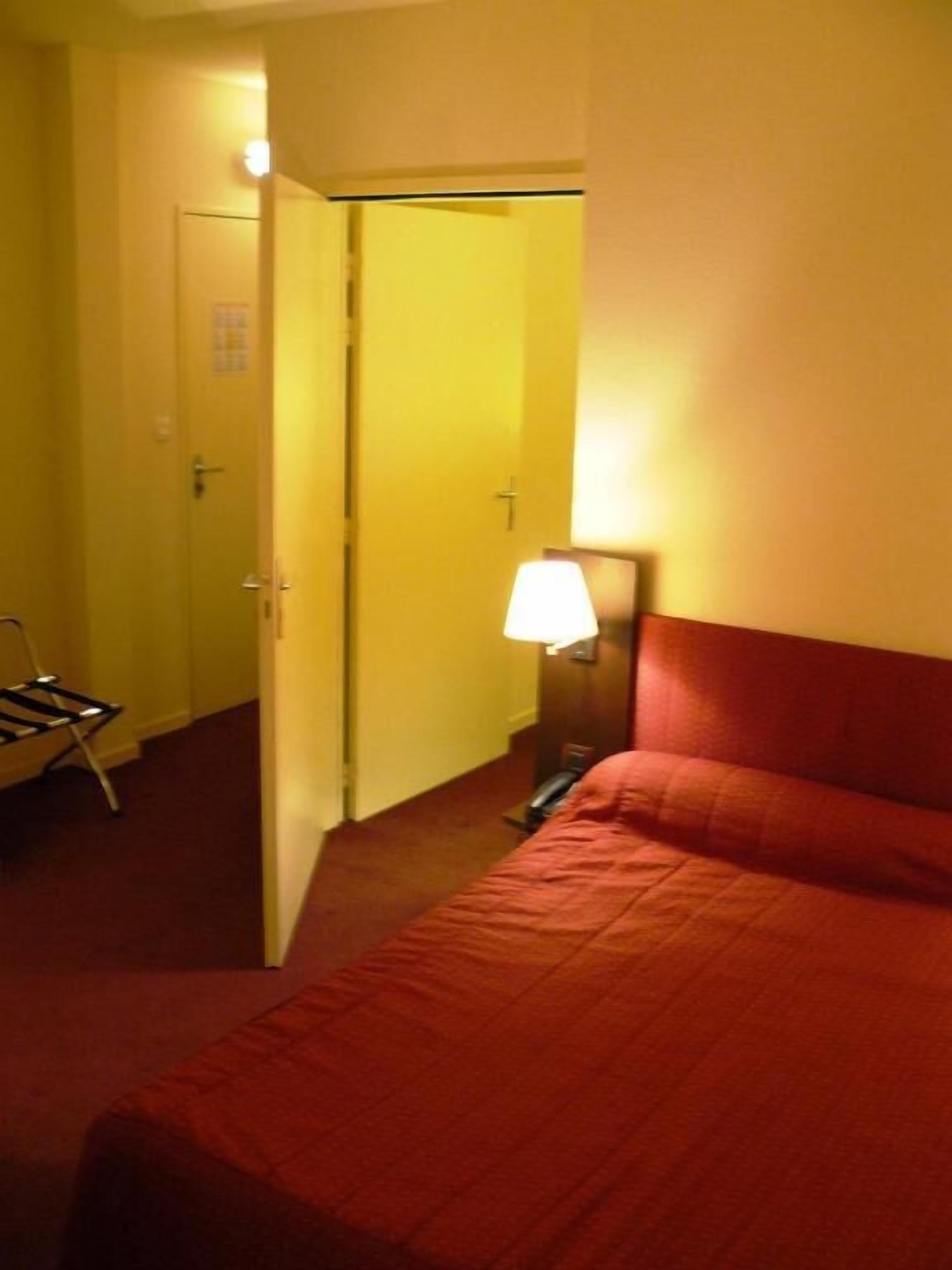A hotel room with a bed, a nightstand, a lamp, and a door leading to another room.