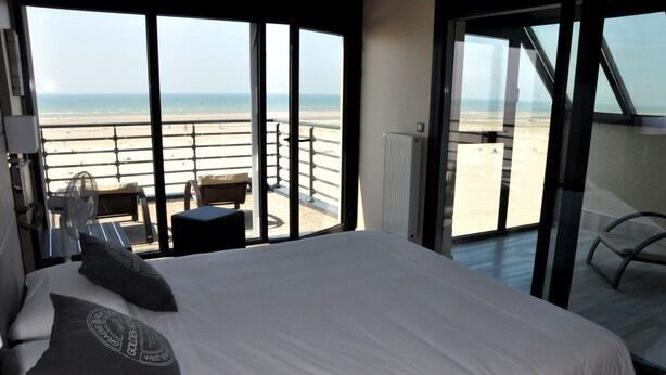 Een hotelkamer met een groot bed, twee kussens, een nachtkastje en uitzicht op het strand door grote ramen.