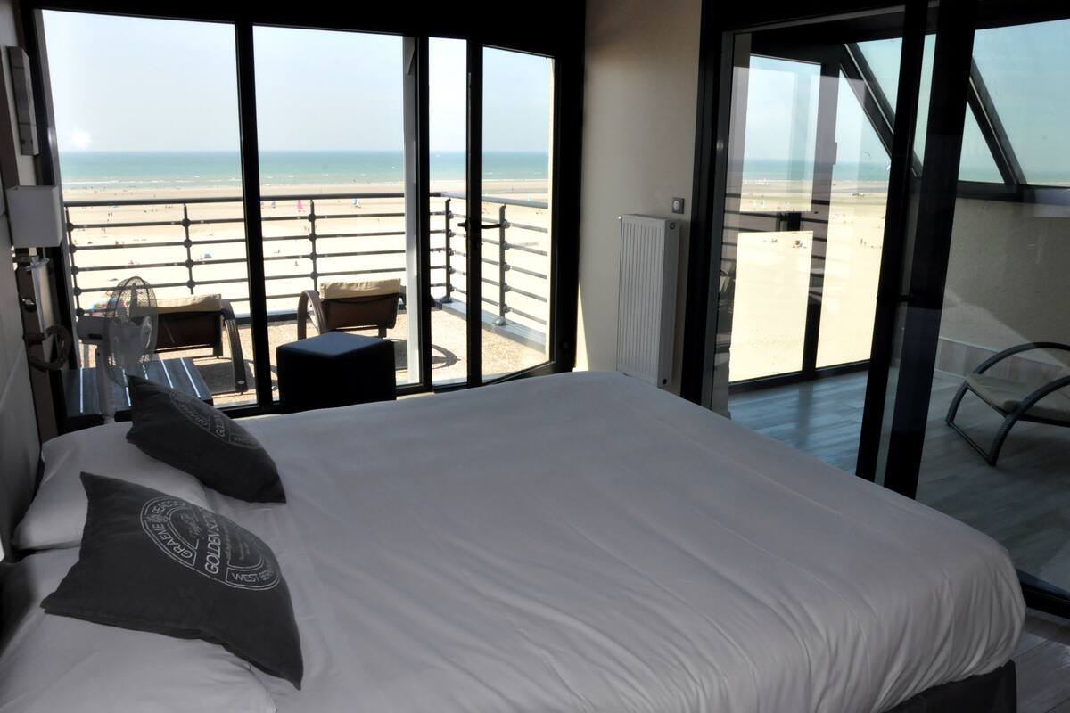 Ein Hotelzimmer mit einem großen Bett, zwei Kissen, einem Nachttisch und Blick auf den Strand durch große Fenster.