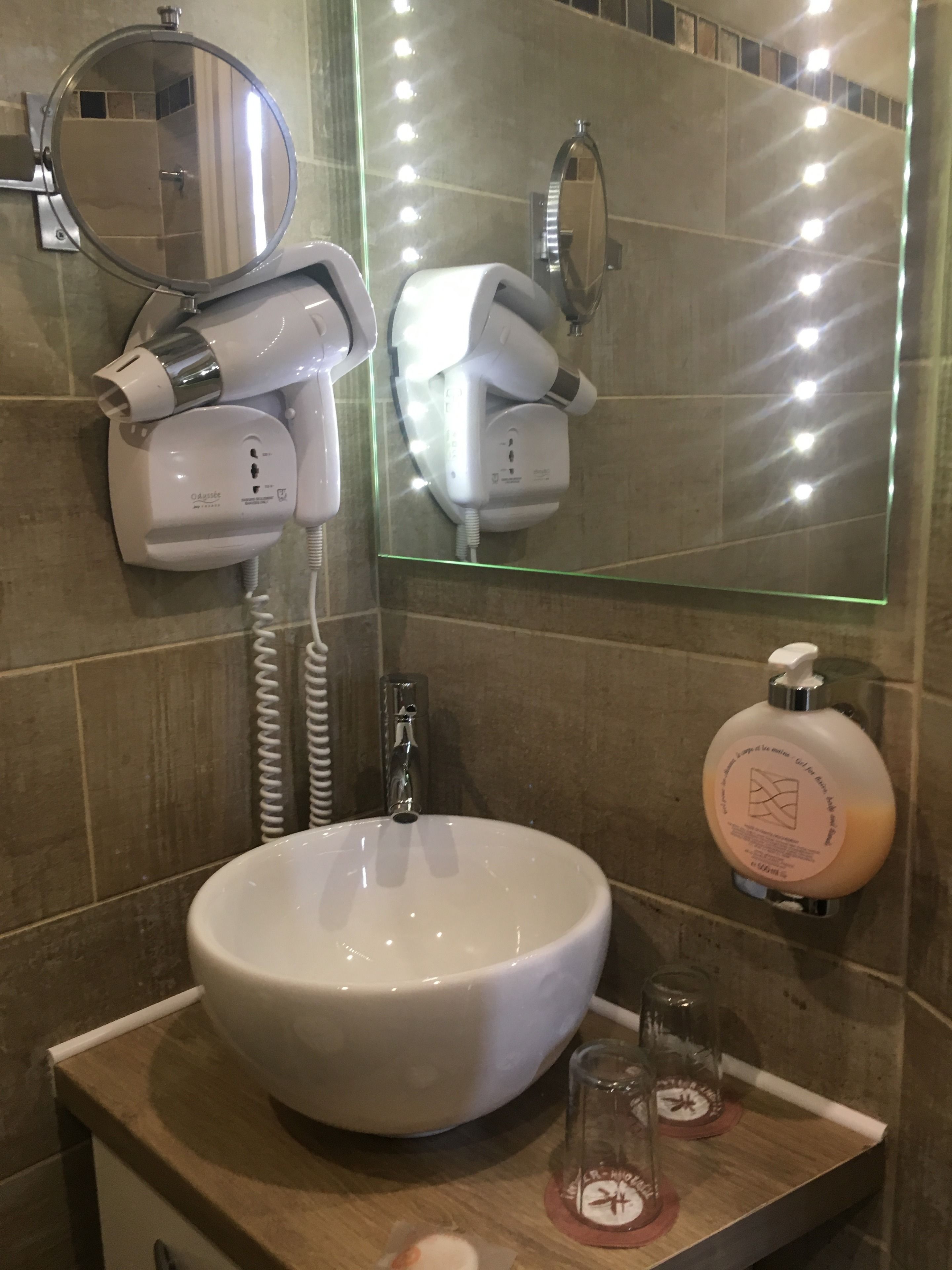 Ein Badezimmer mit einem weißen Waschbecken, einem Spiegel, einem Föhn und einer Shampoo-Flasche.