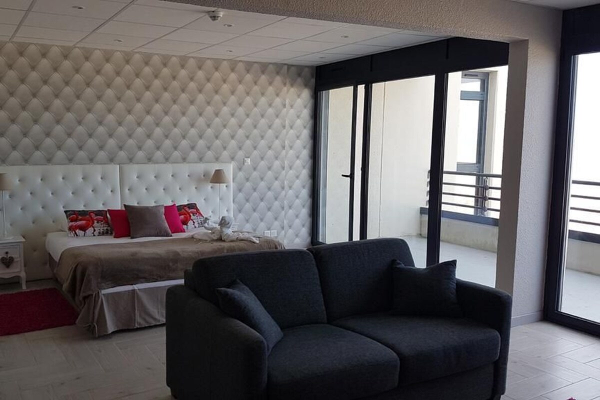 Un dormitorio con cabecera capitonada, cama con almohadas, mesita de noche, alfombra roja y sofá de color oscuro.