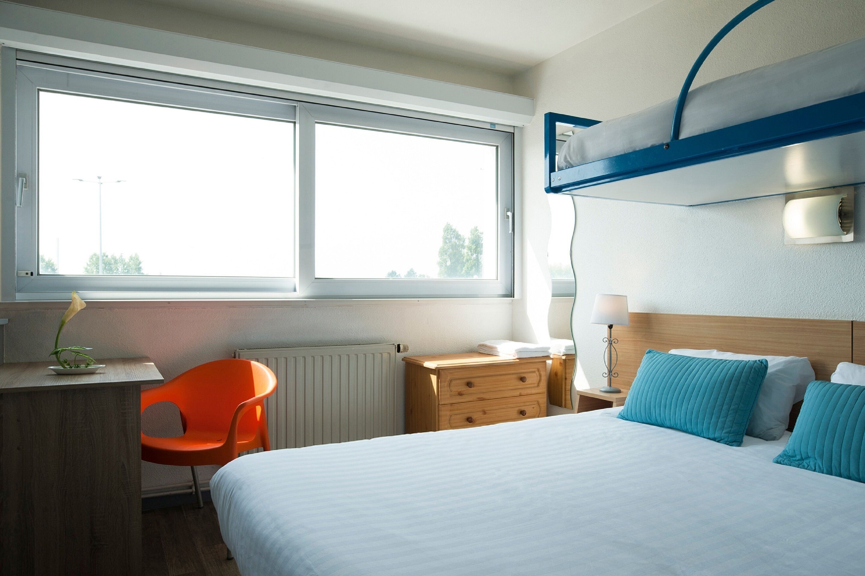 Une chambre d’hôtel avec un lit, un bureau, une chaise orange, une commode et une fenêtre donnant sur un beau paysage.