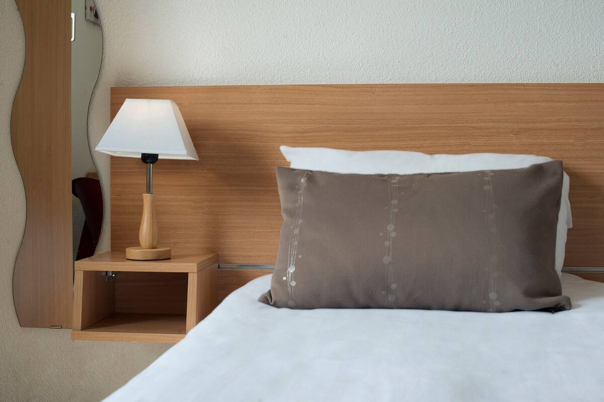 Ein ordentlich bezogenes Bett mit einem weißen und einem braunen Kopfkissen, ein Nachttisch aus Holz mit einer Lampe sowie ein hölzernes Kopfteil.