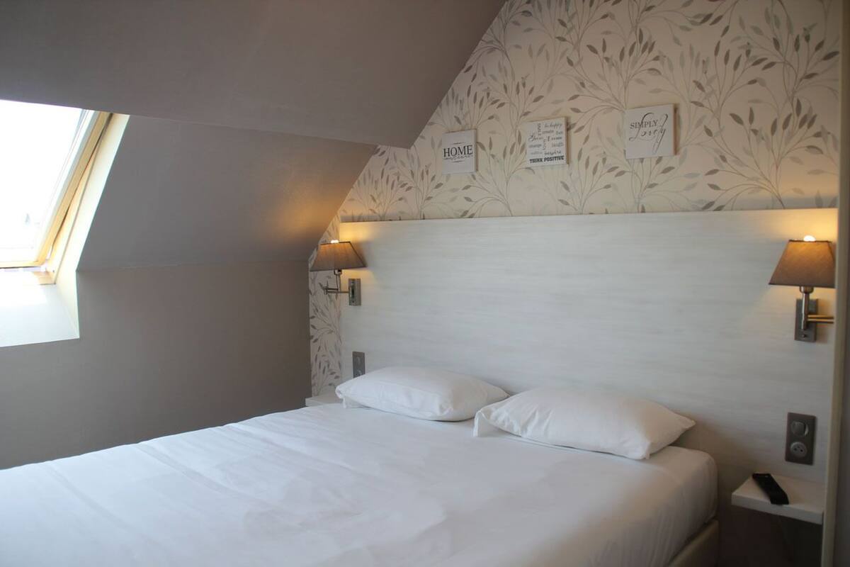 Ein Schlafzimmer mit schrägen Wänden, einem Bett mit weißer Bettwäsche, zwei Nachttischlampen und einem Fenster, das für natürliches Licht sorgt.
