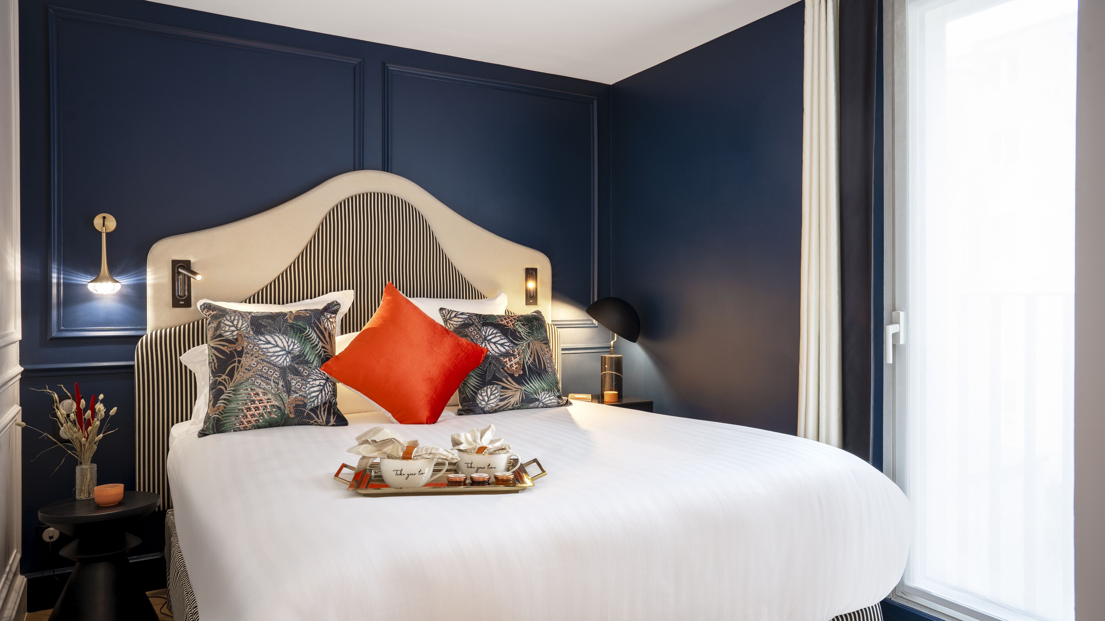 Un lit bien fait, avec un plateau de petit-déjeuner, une table de chevet avec une lampe et une vue sur l’extérieur par la fenêtre.