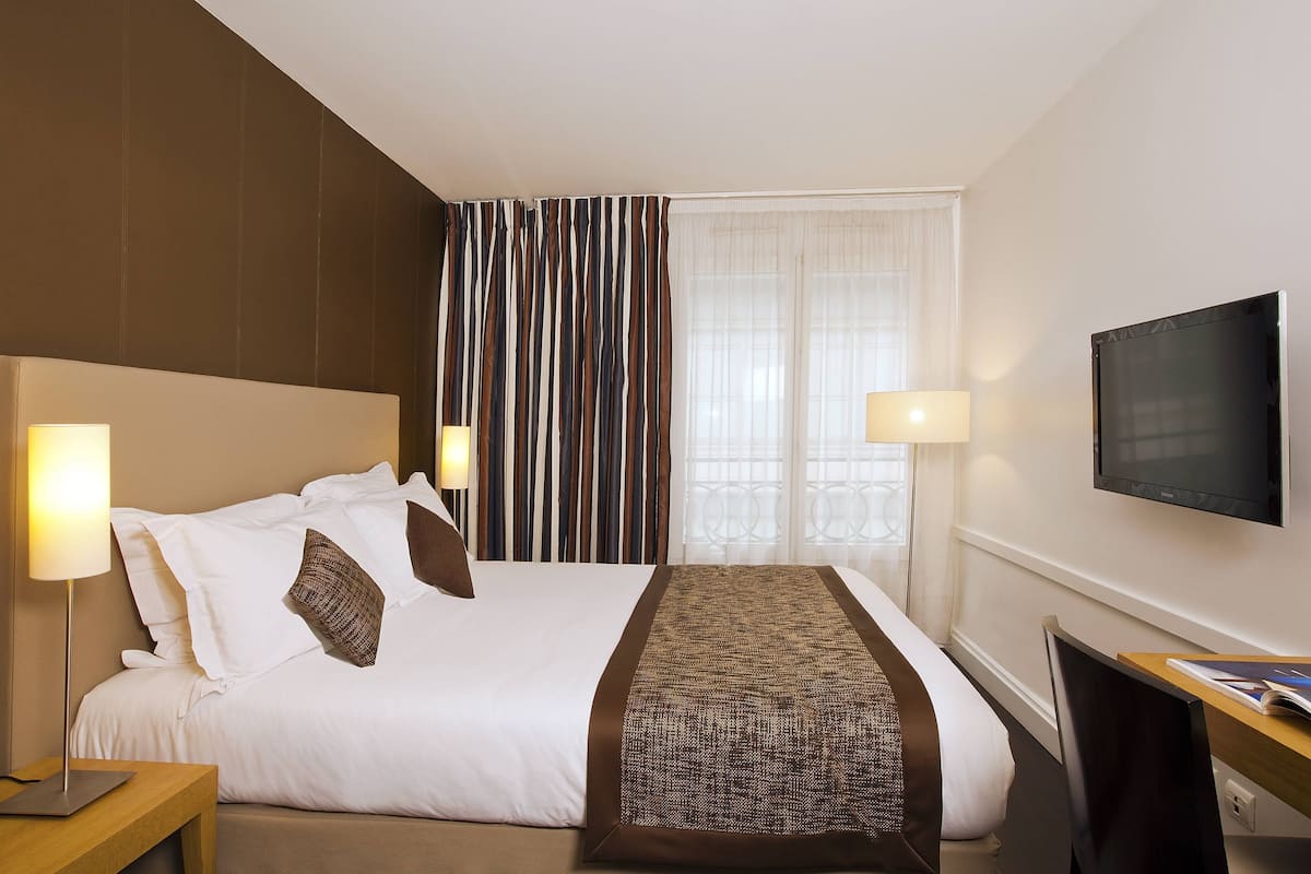 Une chambre d’hôtel avec un lit, deux lampes de chevet, une télévision et une fenêtre avec des rideaux.