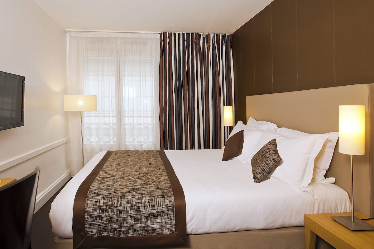 Une chambre d’hôtel avec un grand lit, deux lampes de chevet, un téléviseur fixé au mur et une fenêtre avec des rideaux.