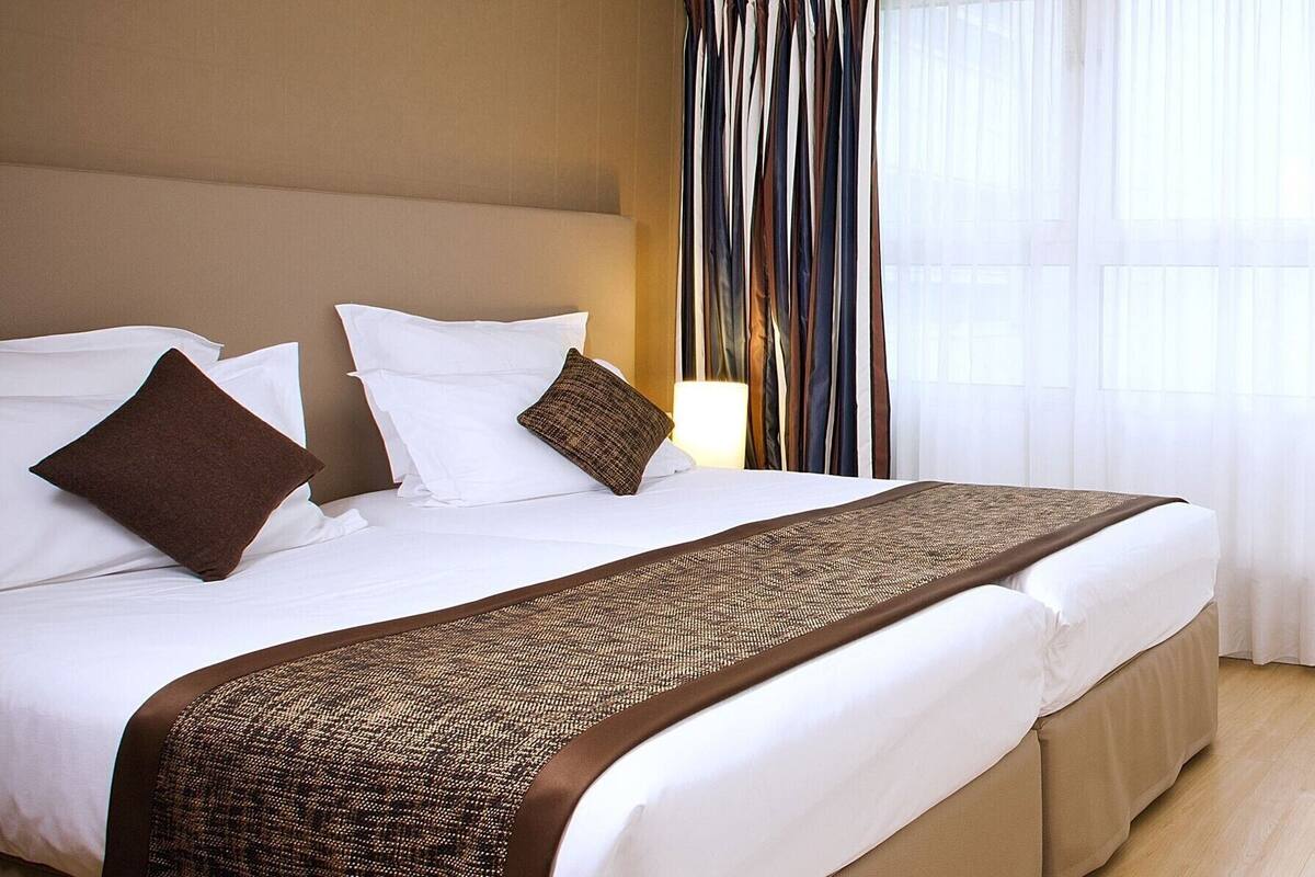 Une chambre d’hôtel avec un grand lit, du linge de lit blanc, un plaid marron et une fenêtre ornée de rideaux légers.