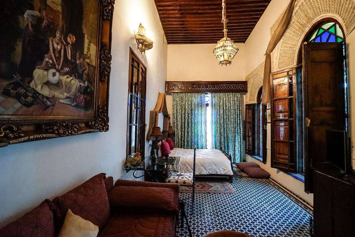 A room with a bed, a sofa, a chandelier, and a large painting.