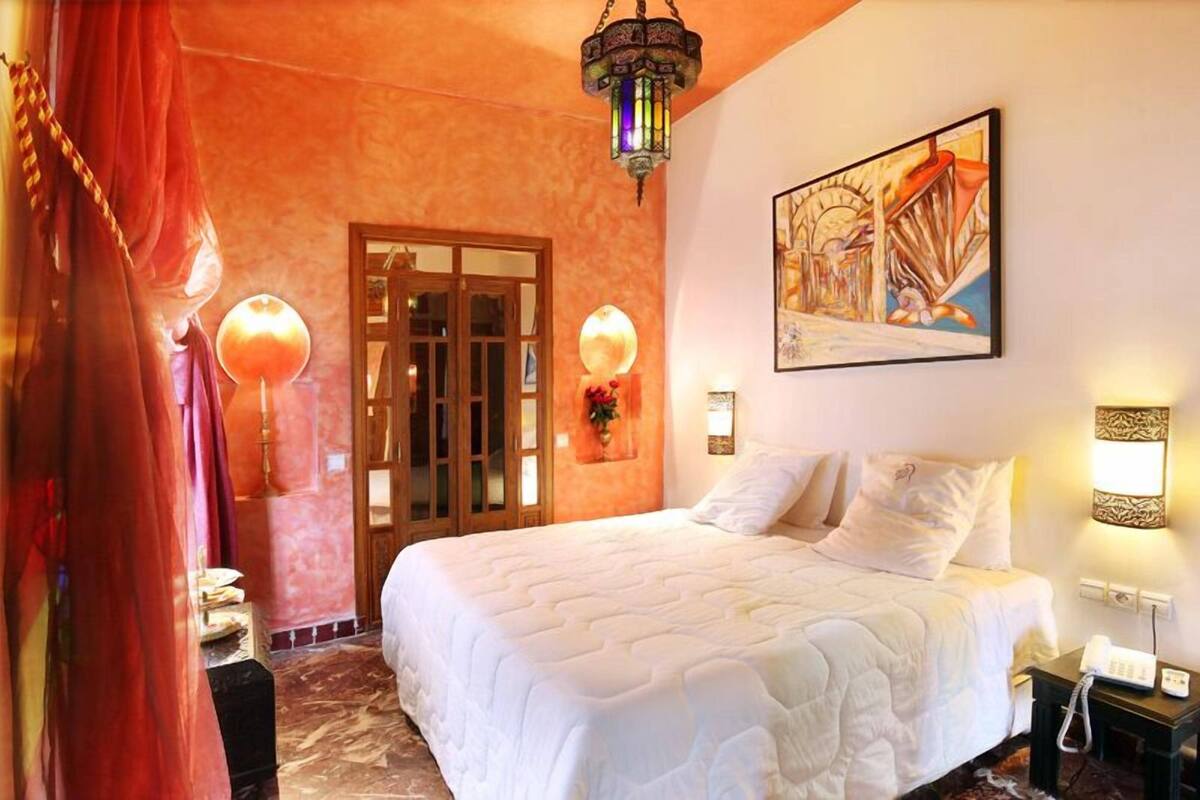A hotel room with a bed, a door, a wall-mounted lamp, and a painting.