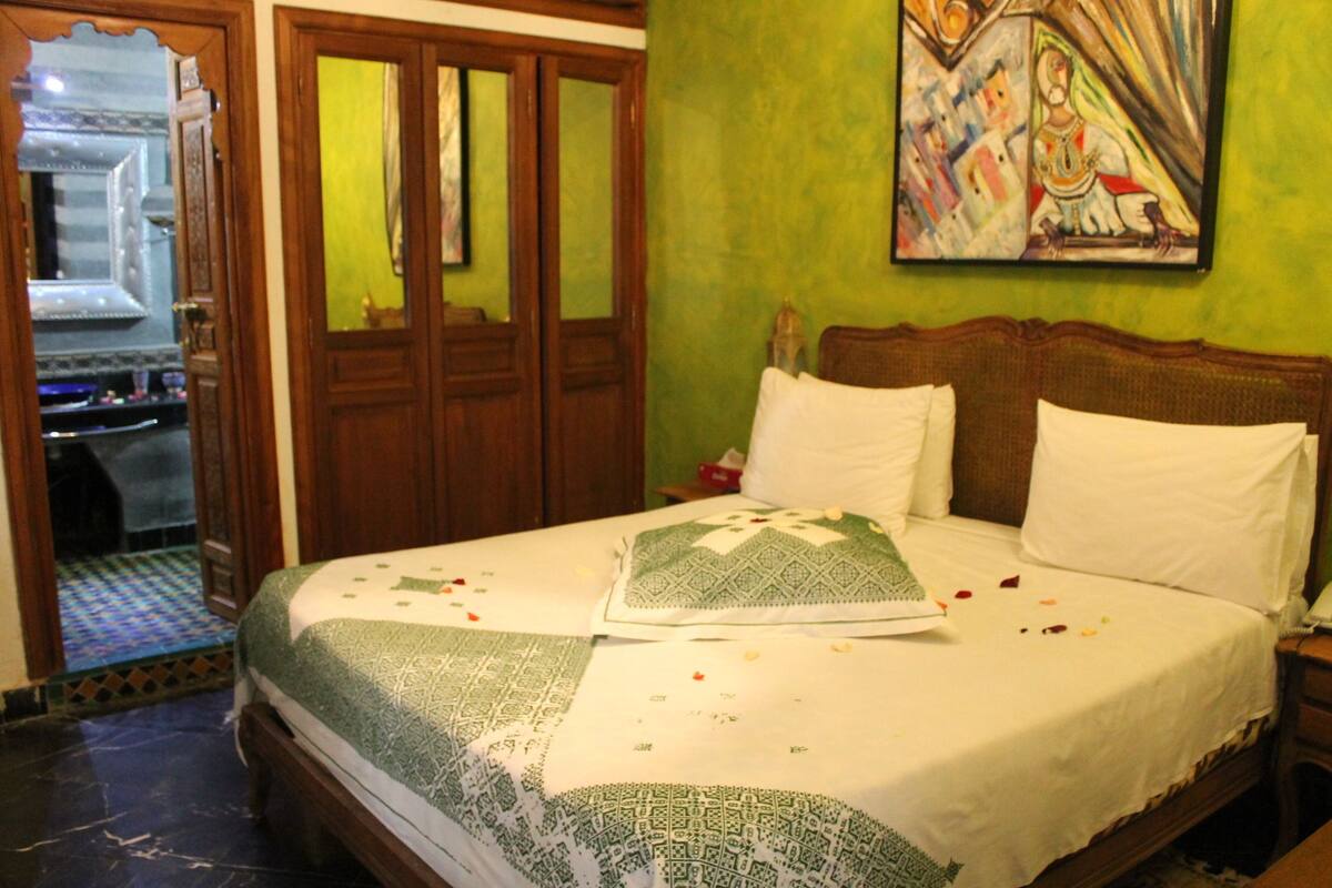A neatly made bed with a patterned bedspread, a green wall with a painting, and a wooden wardrobe with mirrored doors.