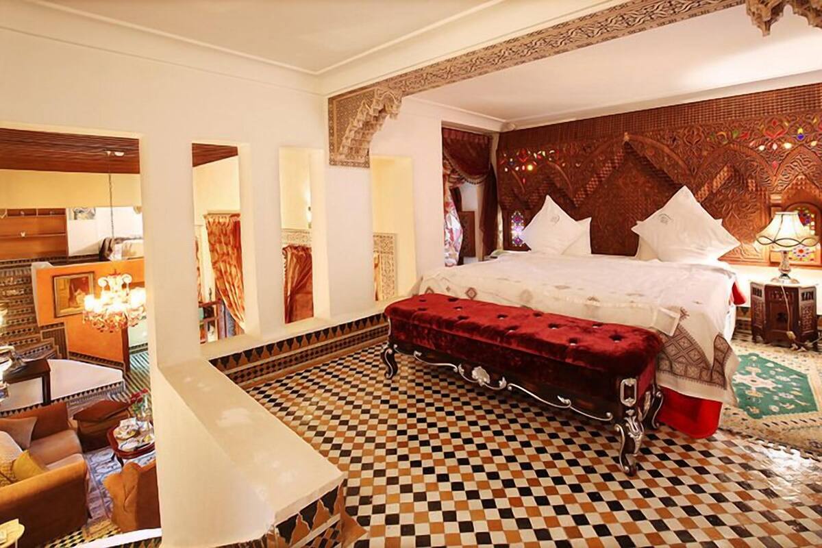 A hotel room with a checkered floor, a large bed with white linens, a red velvet bench, and a decorative headboard.