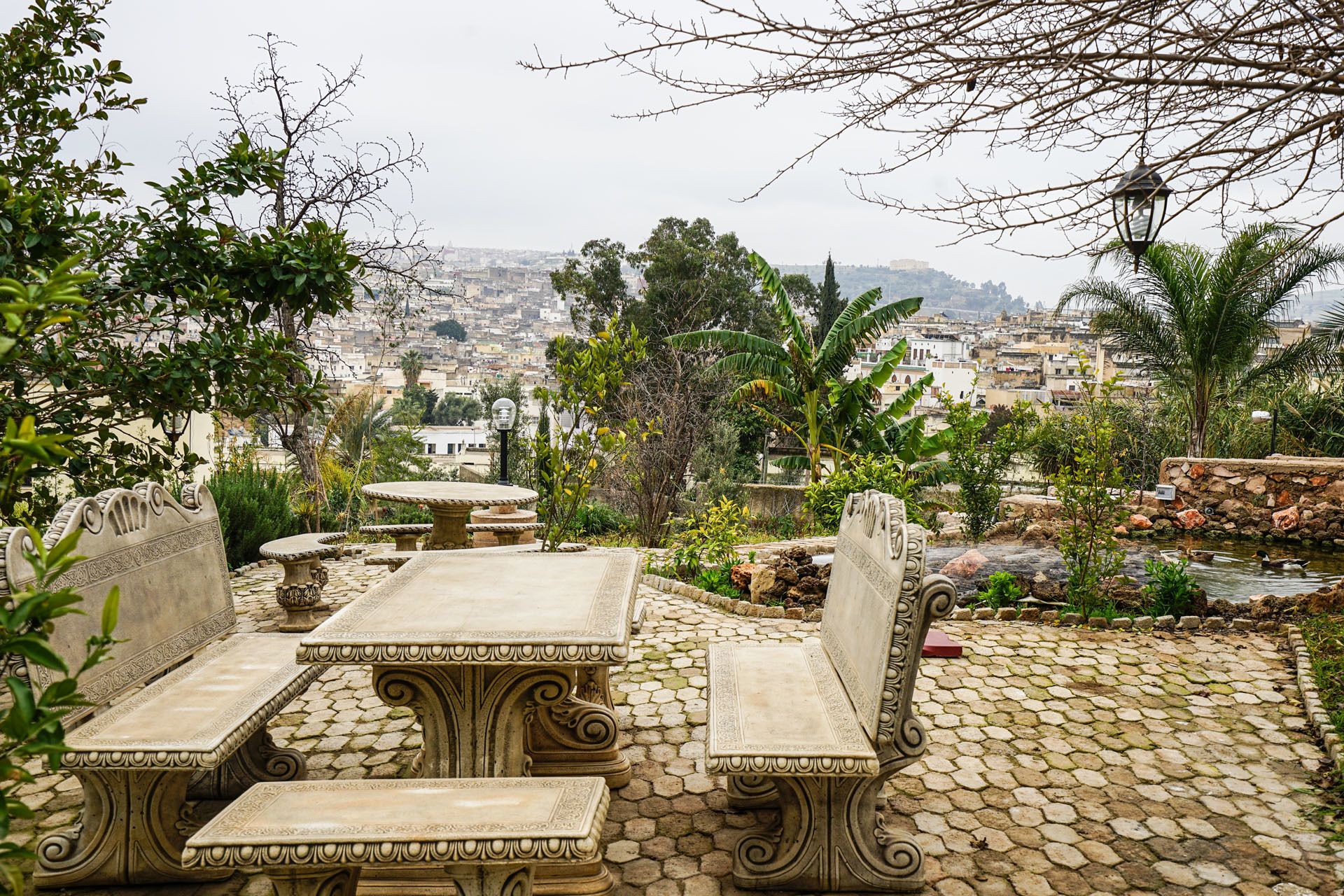 An outdoor seating area with stone tables and benches, surrounded by greenery and overlooking a cityscape.