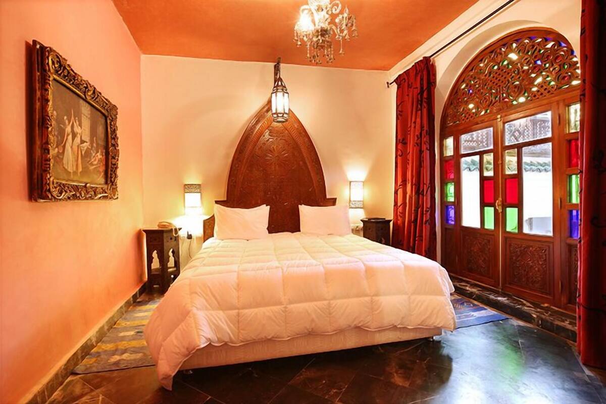A bedroom with a large bed, a chandelier, a decorative arched window, and a framed picture on the wall.