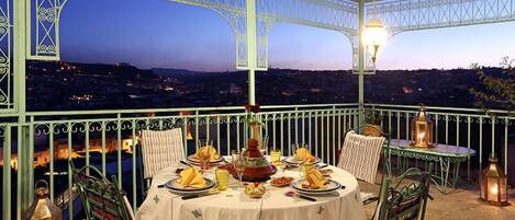 A terrace with a table set for dining, overlooking a cityscape at dusk.