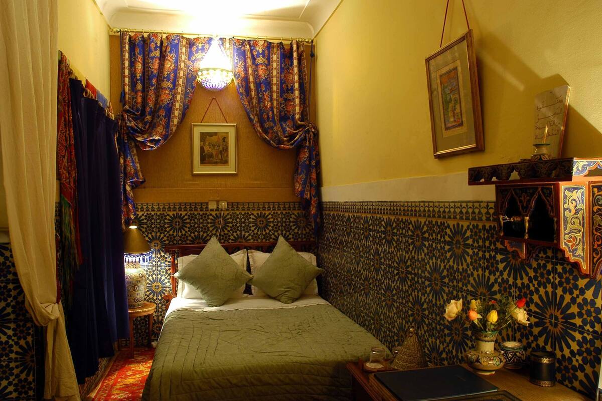 A room with a bed, patterned walls, a lamp, and a vase with flowers.