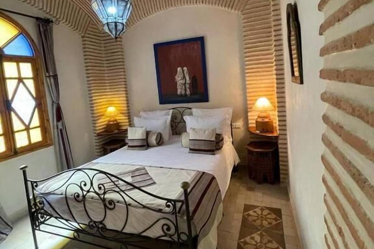 Un dormitorio con cama, mesita de noche, lámpara de pared, un cuadro y una ventana con cortinas.