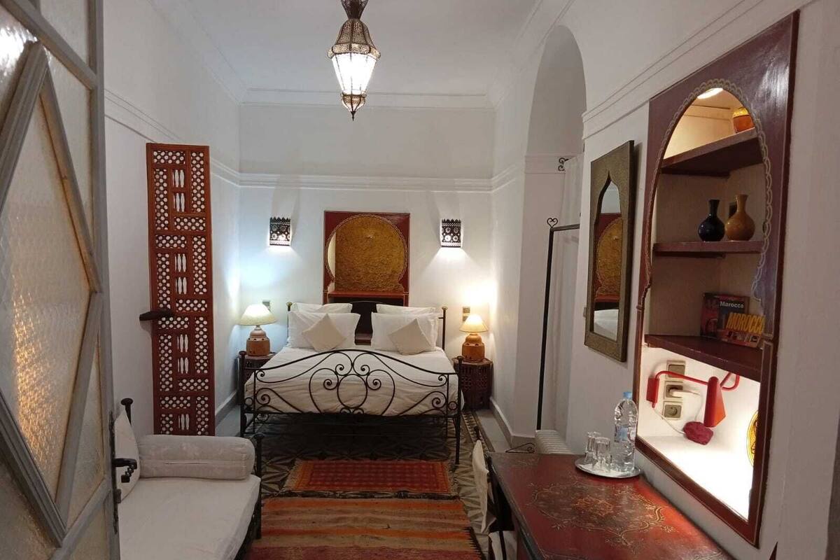 Una habitación estrecha con una cama, una mesita, un espejo y un panel decorativo en la pared.
