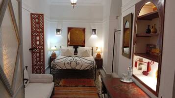 Una habitación estrecha con una cama, una mesita, un espejo y un panel decorativo en la pared.