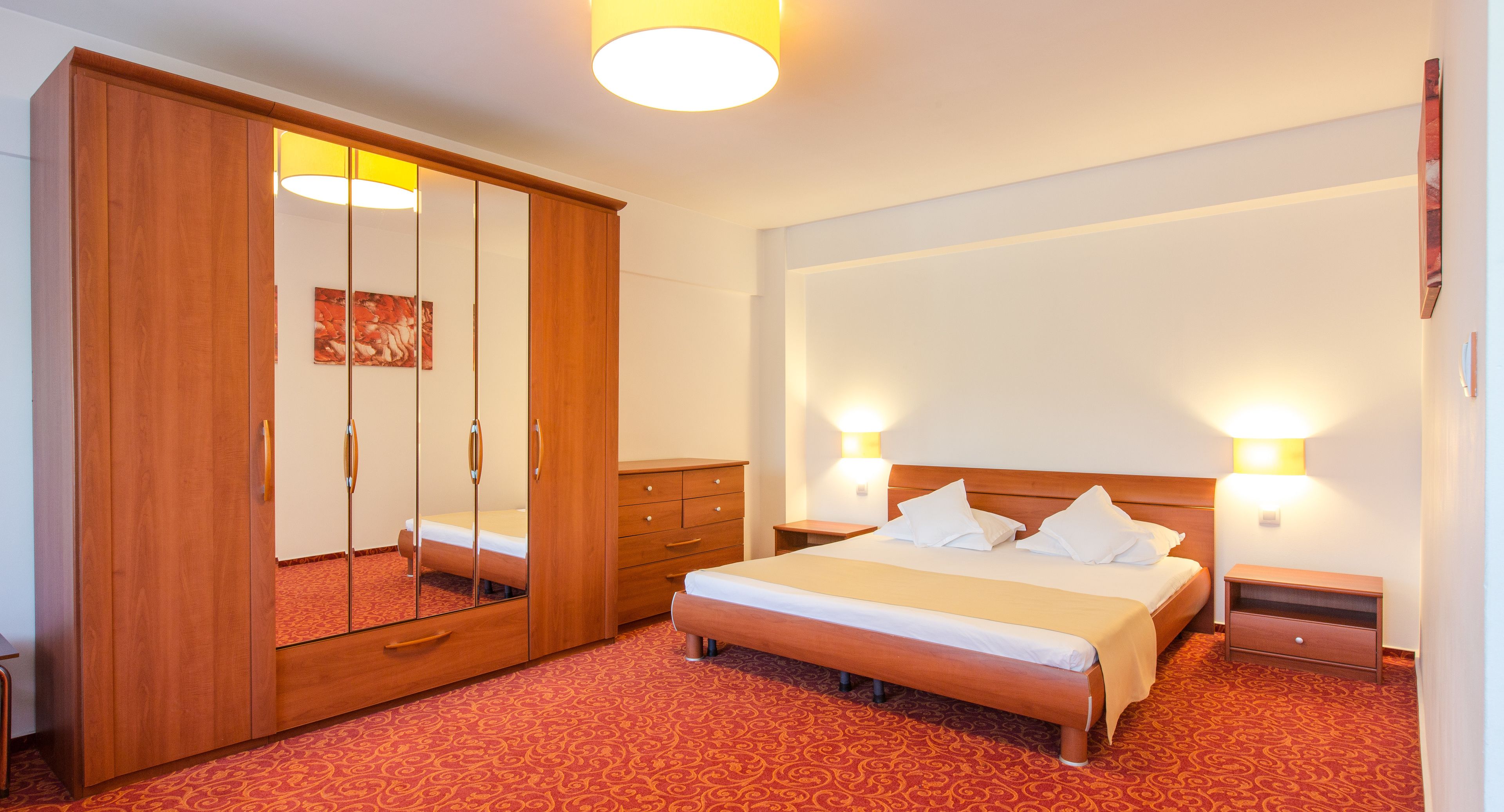 Ein Hotelzimmer mit großem Kleiderschrank, einem Bett mit weißer Bettwäsche, zwei Nachttischen und einem roten Teppich mit Muster.