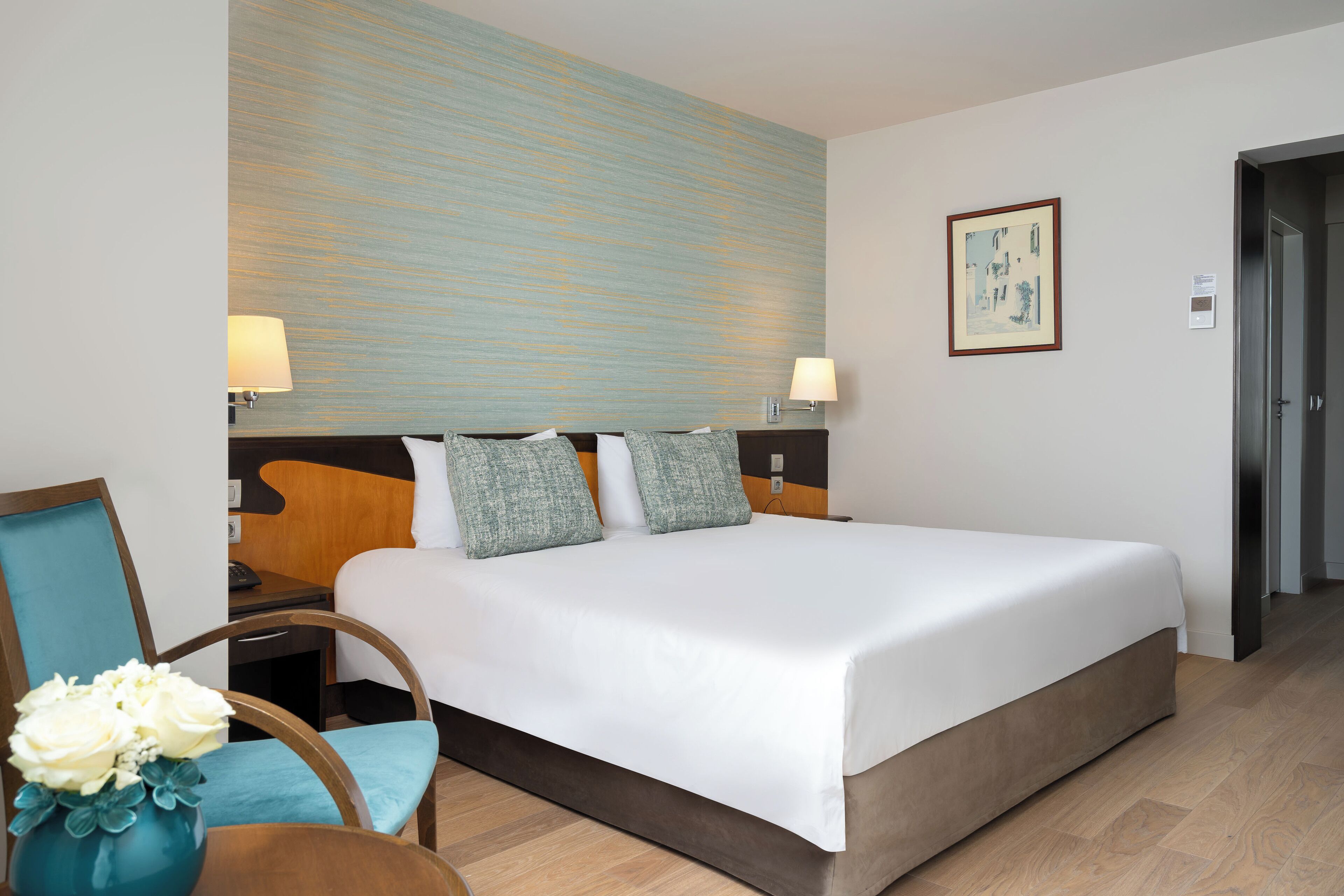 A hotel room with a large bed, two bedside tables with lamps, a chair with a vase of flowers, and a painting on the wall.