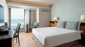 A hotel room with a large bed, a desk with a chair, a small table with a chair, and a view of the ocean through a large window.